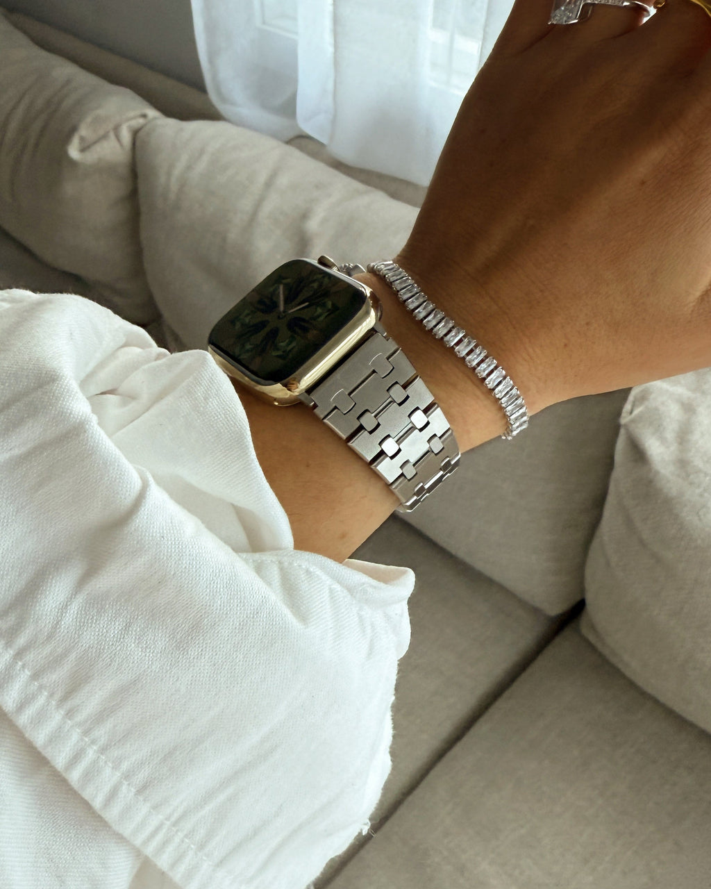 Person wearing a smartwatch and bracelet on a light-colored couch with white curtains in the background.