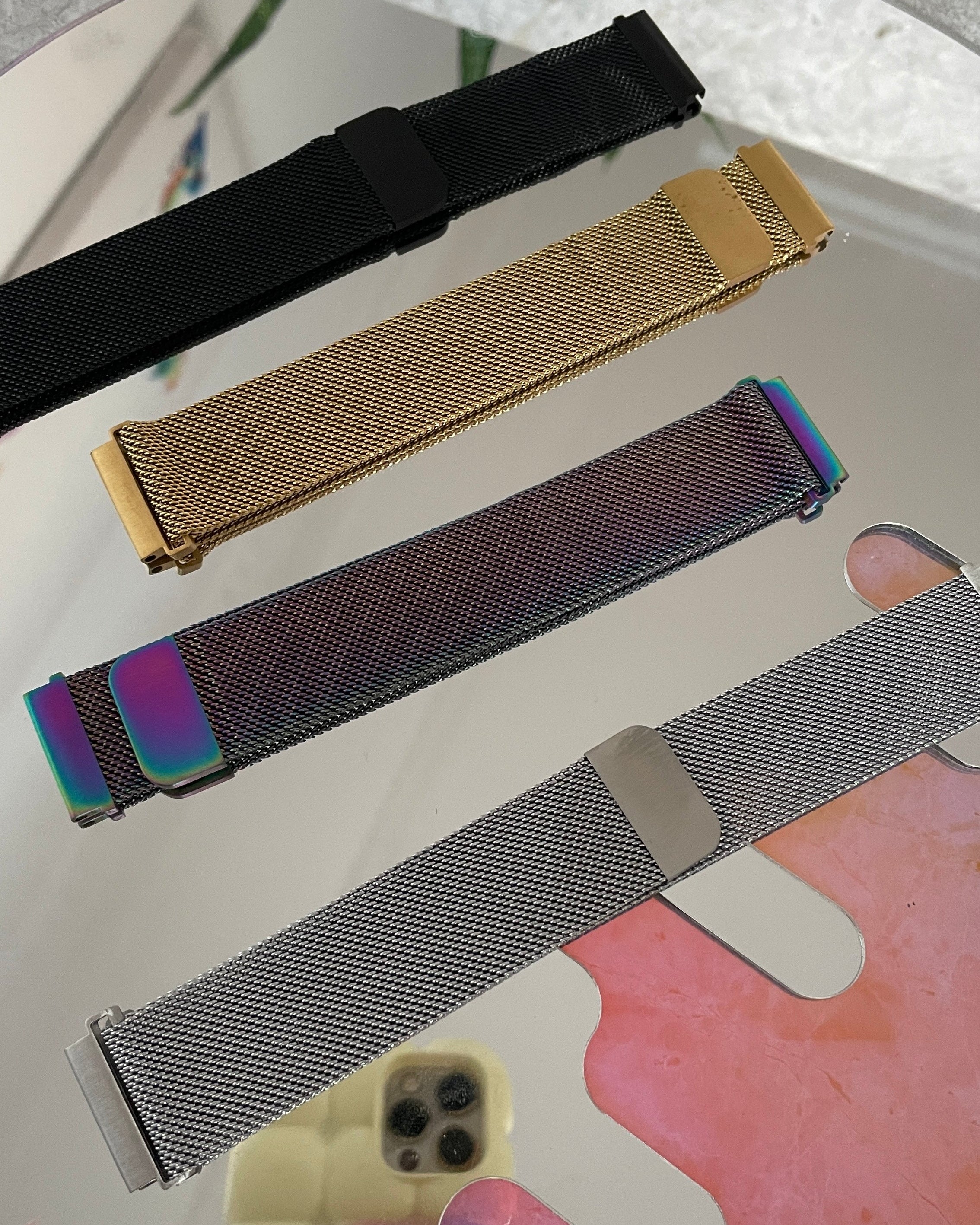 Four mesh watch straps in black, gold, rainbow, and silver on a reflective surface.