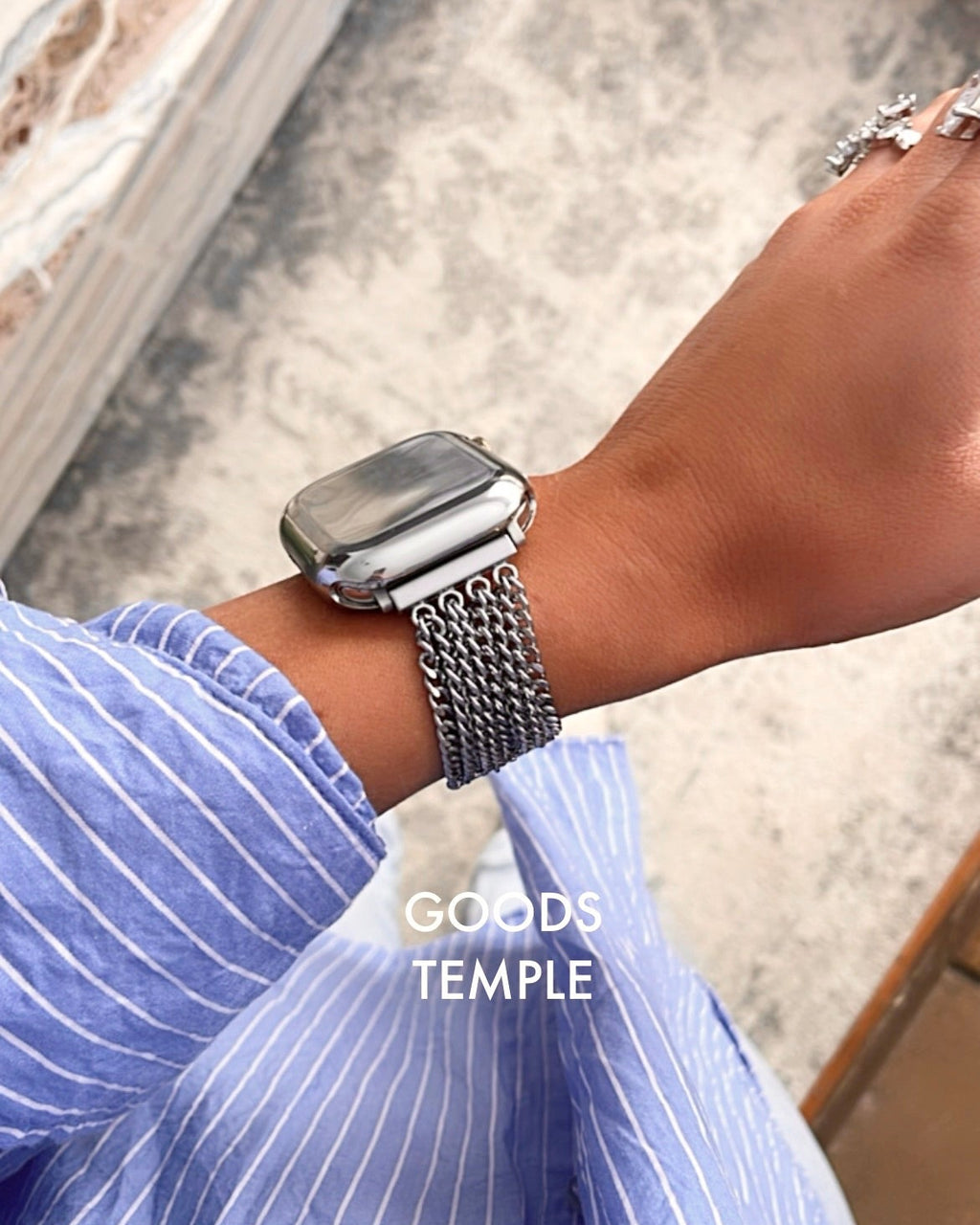 Person wearing a silver watch with a chain strap on a blurred background, featuring the brand 'GOODS TEMPLE'.