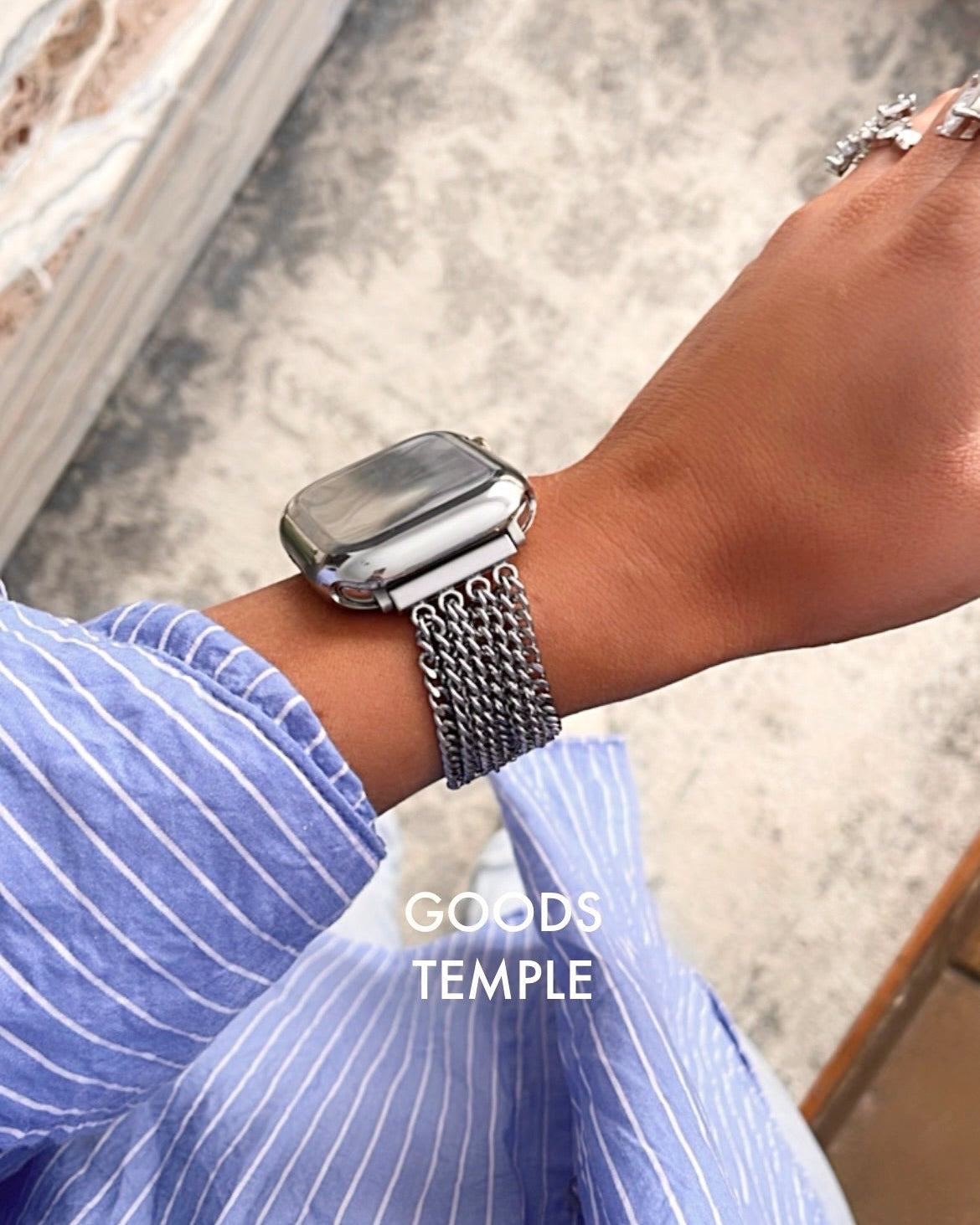 Person wearing a silver watch with a chain strap on a blurred background, featuring the brand 'GOODS TEMPLE'.