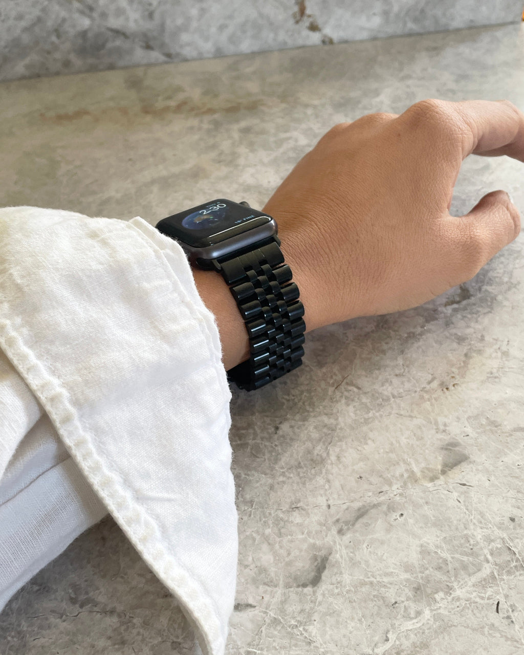 Hand wearing a black jubilee date smartwatch band on a light stone surface