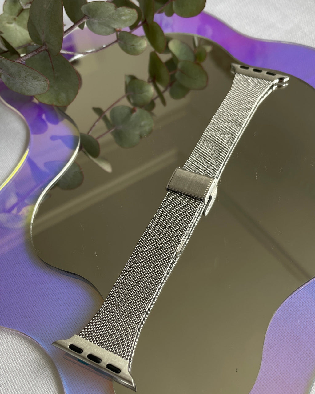 Silver mesh watch band on a reflective surface with greenery in the background