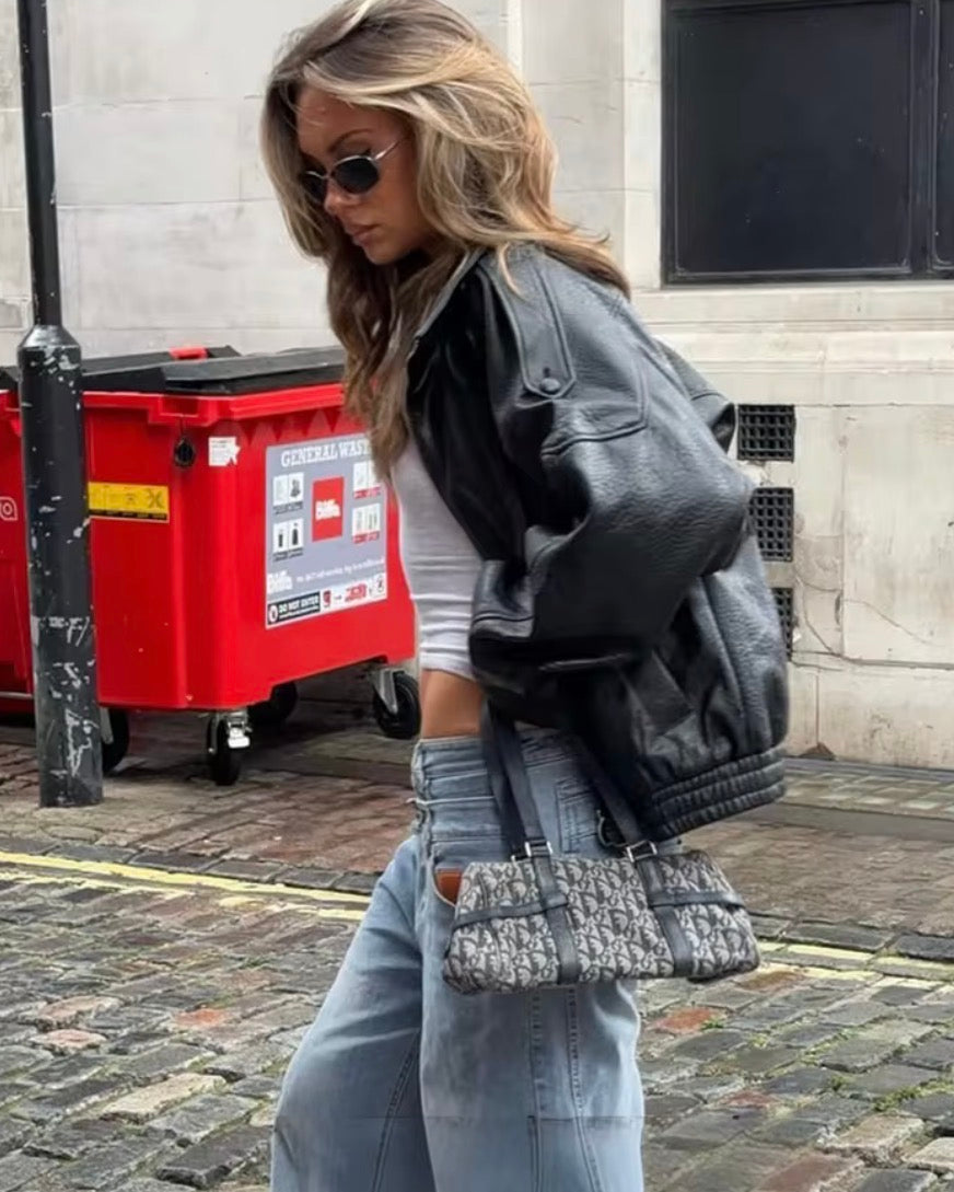 Woman in black leather jacket and sunglasses walking on a street with a red bin in the background