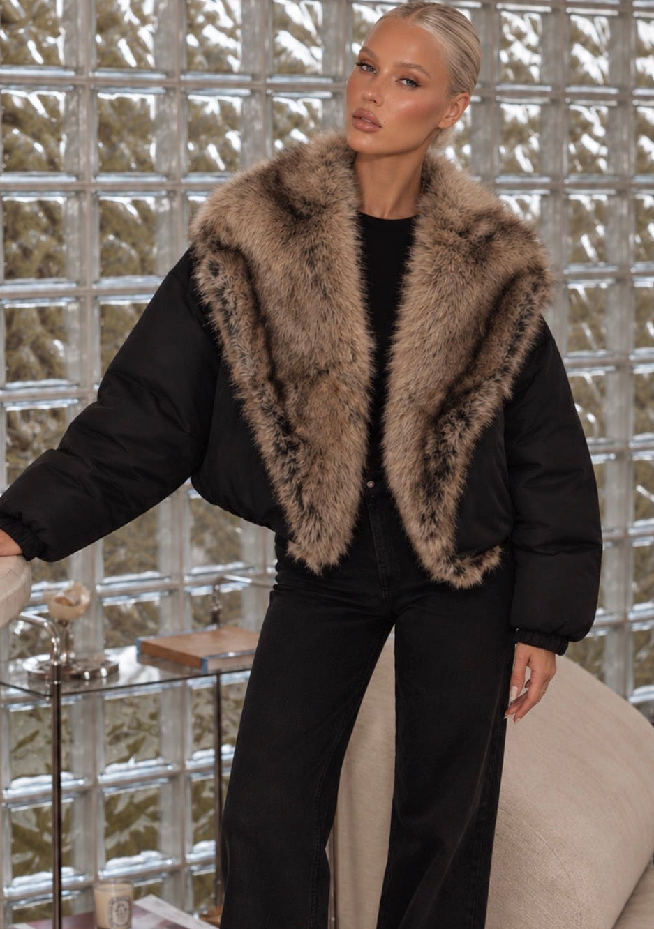 A woman models a black puffer jacket with a large brown and beige faux fur collar, paired with black wide-leg trousers, in an indoor setting with a glass block wall and a beige sofa.