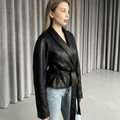 A woman wearing a black wrap-style leather jacket with a matching waist tie and light blue jeans, standing in front of gray curtains.