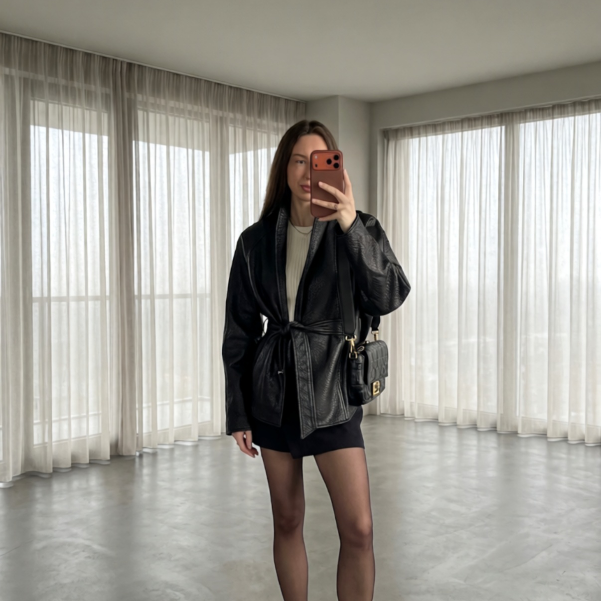 A woman takes a mirror selfie in a minimalist room with sheer white curtains and a gray floor, wearing a black leather wrap jacket, a cream ribbed top, black shorts, and carrying a small black shoulder bag.