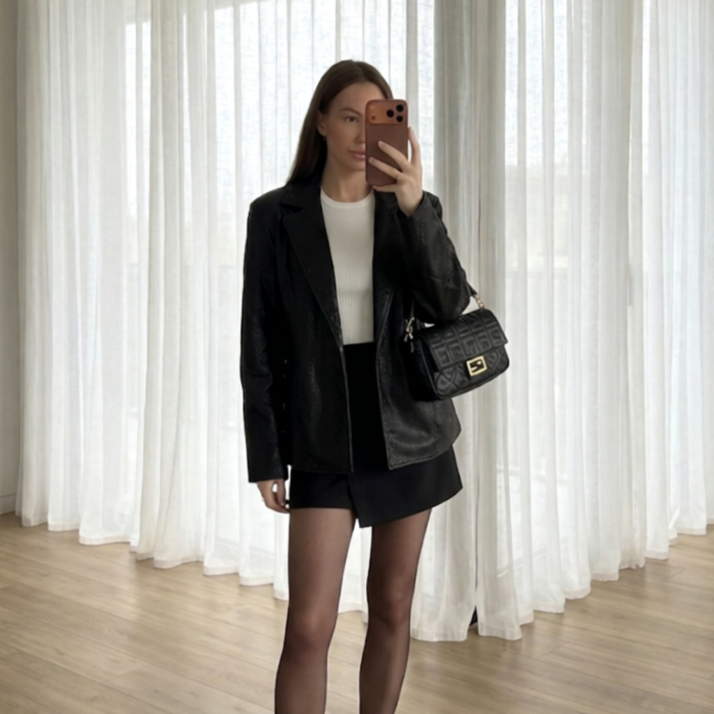 A woman takes a mirror selfie in a room with white sheer curtains and light wood flooring, wearing a black leather blazer, black shorts, a white top, and carrying a black crocodile-embossed shoulder bag.