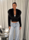 A woman stands indoors wearing a black cropped cardigan with a very large black fur collar, light-wash wide-leg jeans, and black sandals. She is posed in front of white draped curtains with a metallic bowl chair to her left.