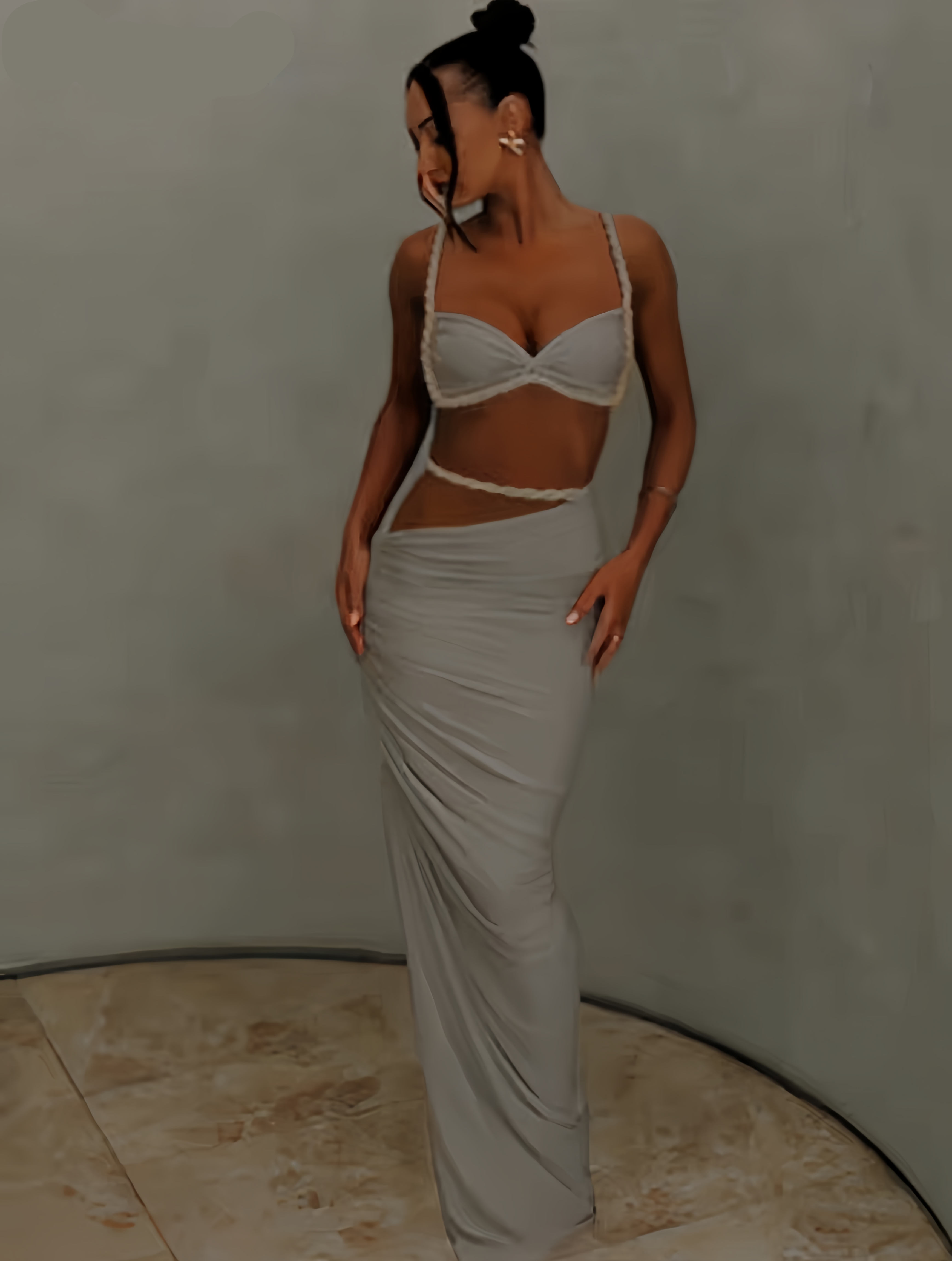 A woman models a light gray two-piece set consisting of a bralette top with rope trim and a matching draped maxi skirt, set against a neutral gray studio backdrop.